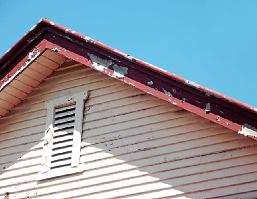 Weathered fascia board with peeling paint above aged soffit at a roof gable